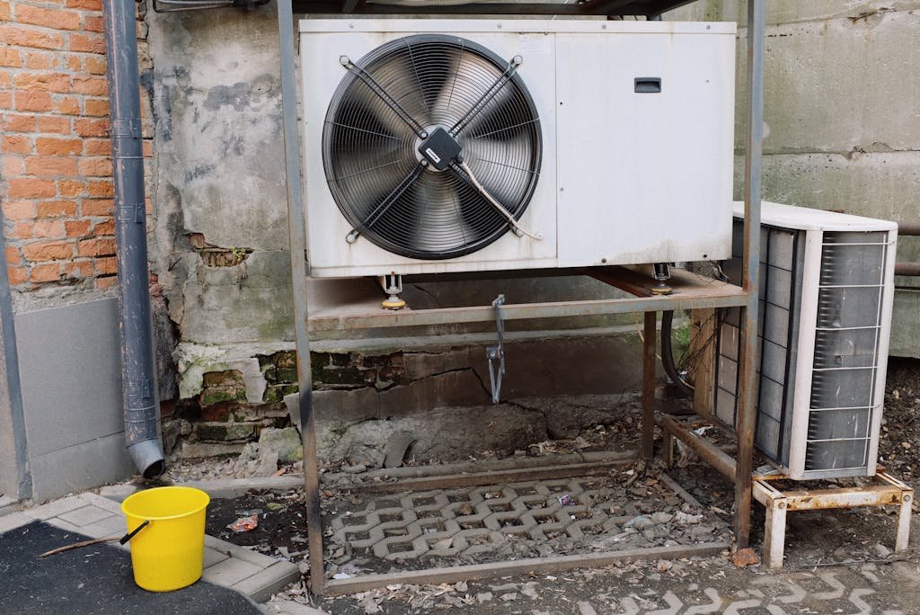 Exterior view of an industrial air conditioning unit with visible wear and rust, set against a shabby wall.